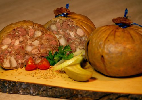 Zwei Blasen Sülzwurst, eine davon halbiert. Garniert mit saurer Gurke, Tomate und Petersilie.