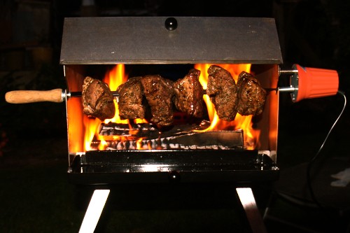 Mutzbratengrill mit sechs Stück Mutzbraten auf einem Spieß über Feuer in der Dunkelheit